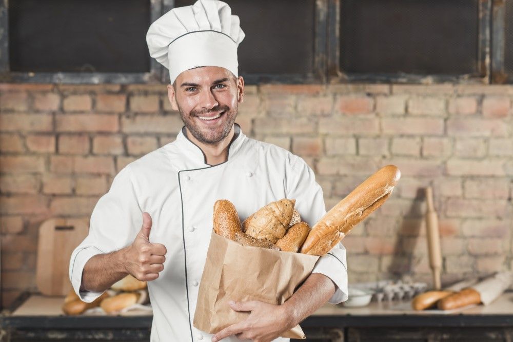 smiling-young-male-baker-holding-loaf-breads-paper-bag-showing-thumb-up-sign