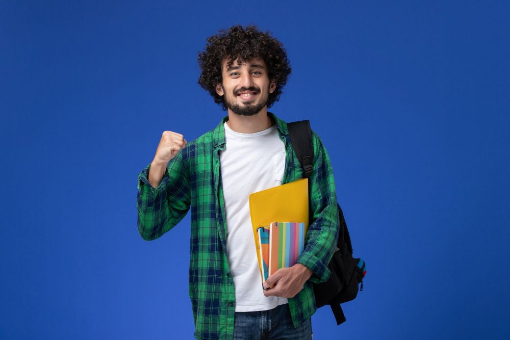 front-view-of-male-student-wearing-black-backpack-holding-copybooks-and-files-on-blue-wall