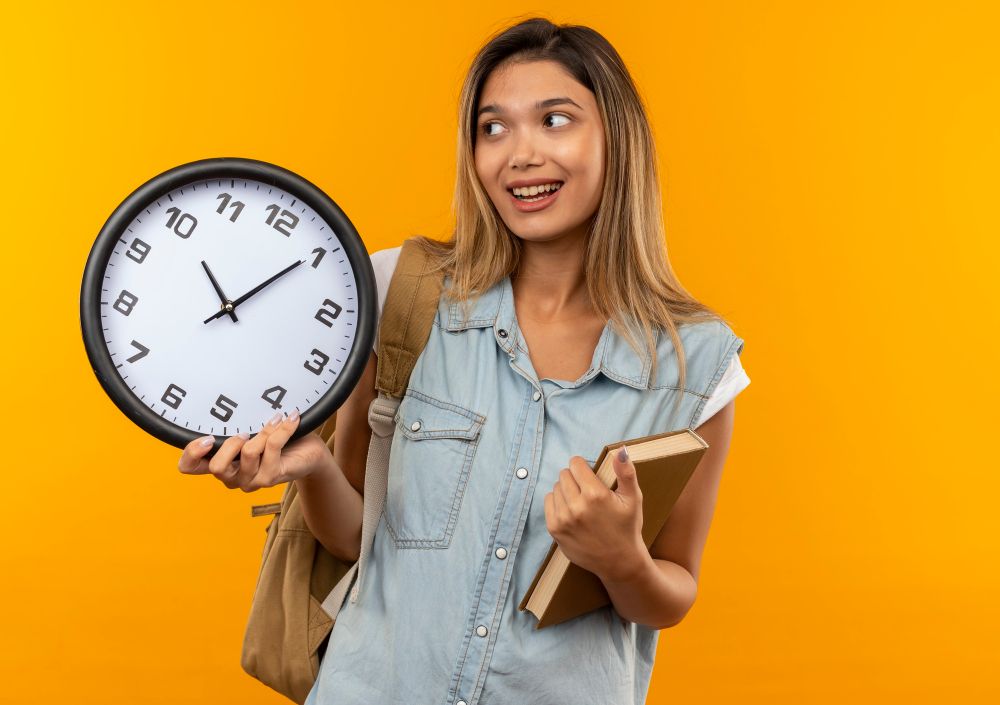 joyful-young-pretty-student-girl-wearing-back-bag-holding-book-and-clock-and-looking-at-side-isolated-on-orange